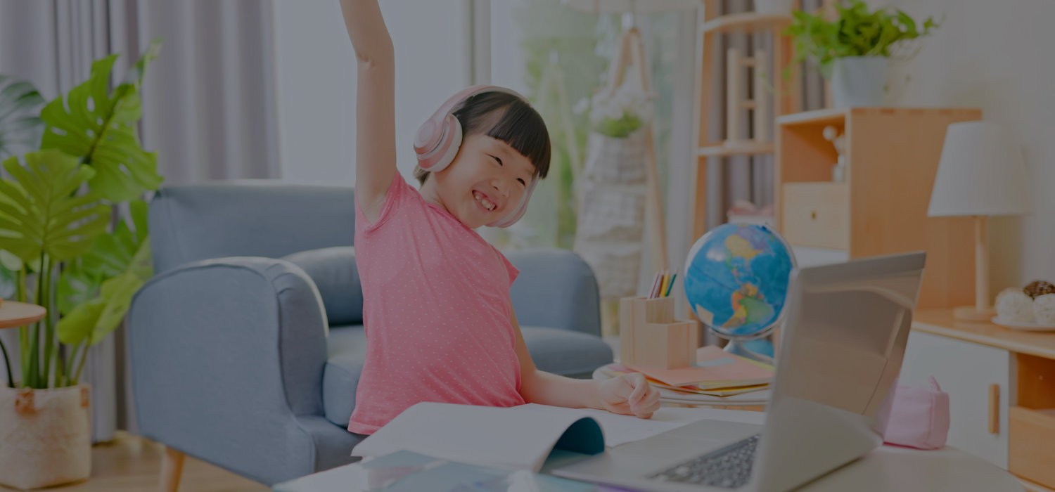 Child raising their hand while on a laptop with a book in front fo her